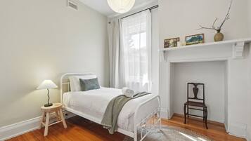 A bedroom with a bed, a small bedside table with a lamp, a chair, a fireplace, and a shelf with pictures and a vase.