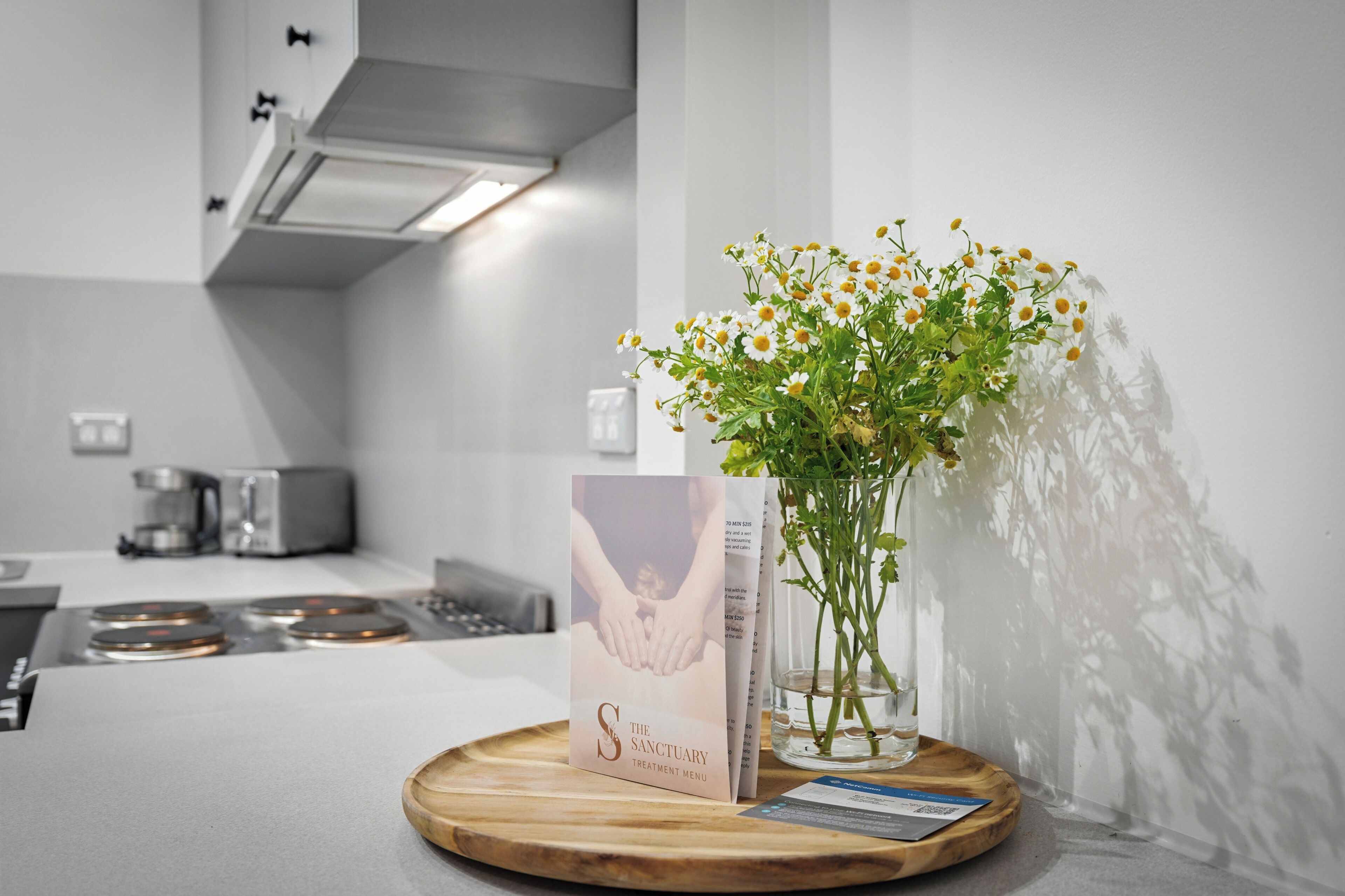Un folleto titulado 'The Sanctuary' sobre una bandeja de madera en una cocina moderna con encimera blanca y electrodomésticos de acero inoxidable.