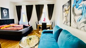A bedroom with a bed, a blue sofa, a dining table, and a painting on the wall. - 1BR Apt for 5P, 10-Min Transport to Stadthalle (Vienna)