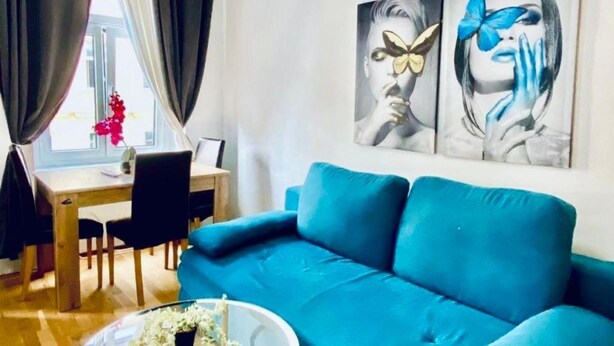A room with a blue sofa, a dining table with chairs, and a window with curtains. - 1BR Apt for 5P, 10-Min Transport to Stadthalle (Vienna)