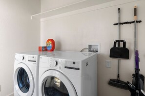 A laundry area with two front-loading washing machines, a bottle of Tide detergent, and a broom and dustpan hanging on a wall-mounted rack.