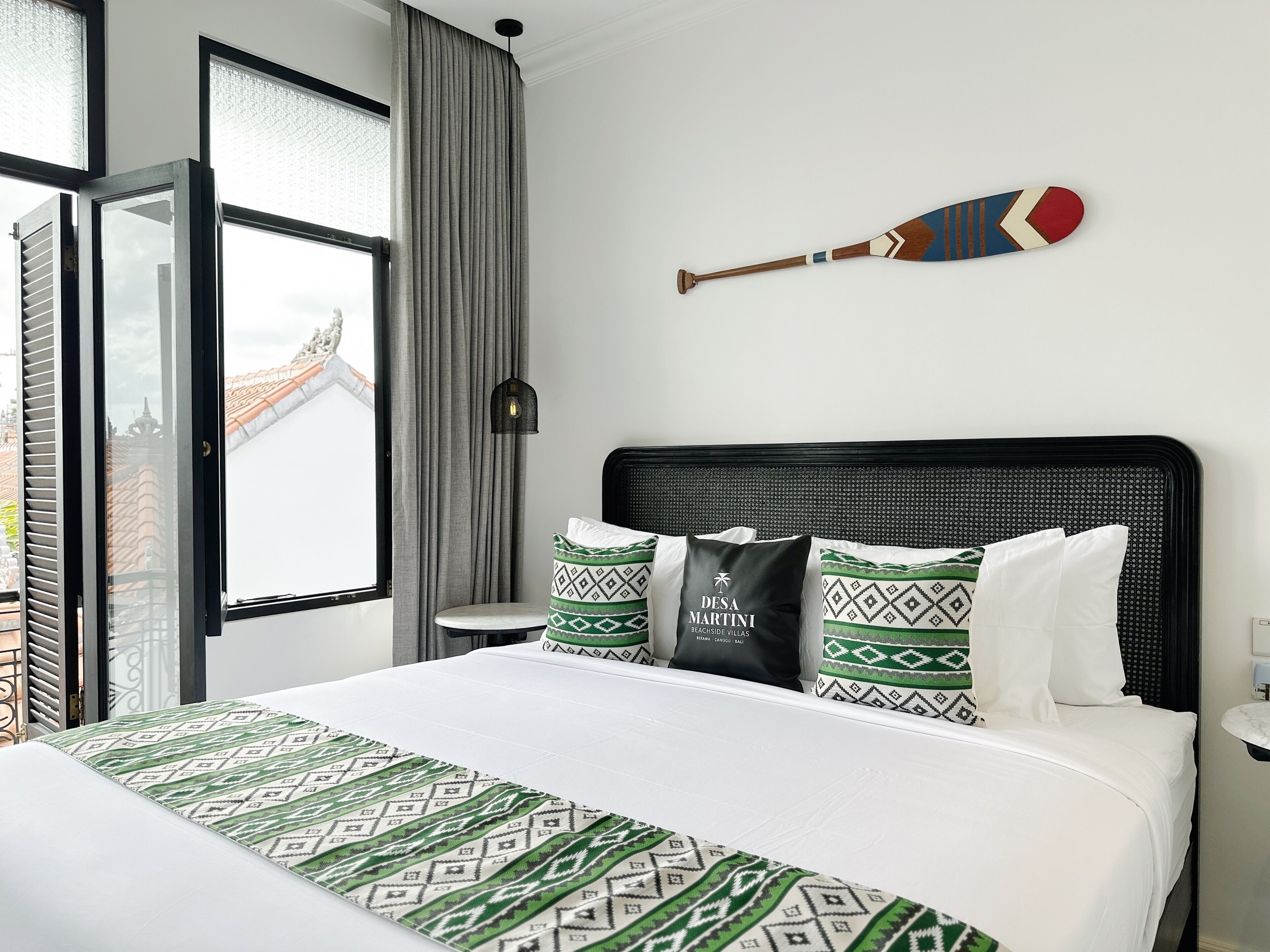 A hotel room with a bed, a view of a building through a window, a bat hanging on the wall, and a lamp.