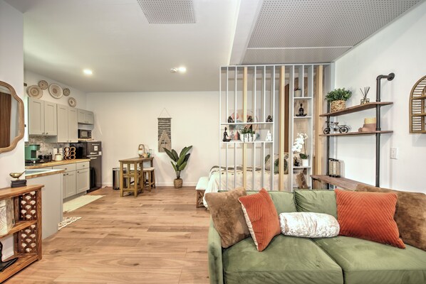 Una sala de estar moderna con un sofá verde, piso de madera, zona de cocina con armarios blancos y una planta decorativa.