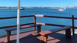 A wooden deck with a white umbrella and benches overlooking a body of water with boats and a distant city skyline. - Aloha home-Amazing location for all GC has to offer amazing home for the family. (Labrador)