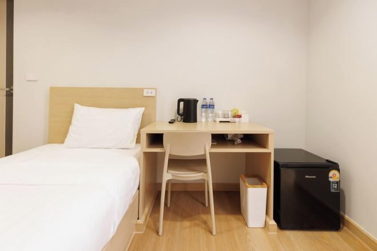 A hotel room with a bed, a desk, a chair, a coffee maker, a refrigerator, and a trash bin.
