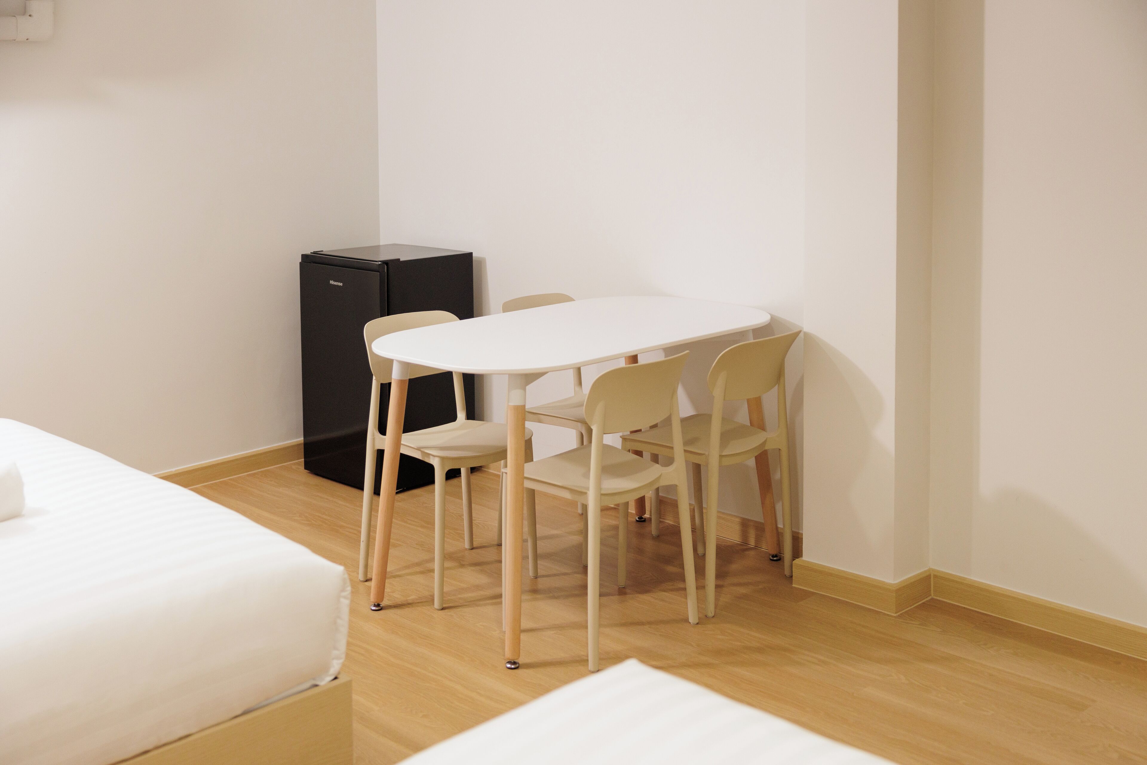 Une petite pièce propre, avec un lit blanc, une table blanche et quatre chaises. Un meuble noir est adossé au mur.