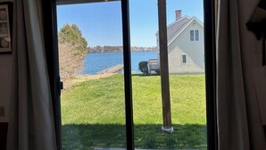 A view through a glass door to a grassy yard with a wooden deck, a house, and a body of water. - Enjoy Stunning Ocean Views & Coastal Access at Sea Mist Overlook! (Harpswell)