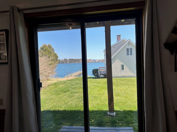 A view through a glass door to a grassy yard with a wooden deck, a house, and a body of water. - Enjoy Stunning Ocean Views & Coastal Access at Sea Mist Overlook! (Harpswell)