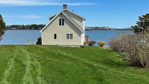 A single-story house with a gabled roof, a chimney, and a small porch with a chair. It is situated on a grassy area with a body of water in the background. - Enjoy Stunning Ocean Views & Coastal Access at Sea Mist Overlook! (Harpswell)