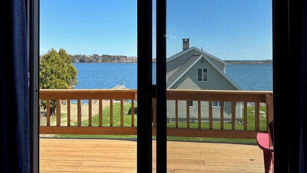 A view from a balcony with a wooden deck overlooking a lake, a house with a gabled roof, and a clear blue sky. - Enjoy Stunning Ocean Views & Coastal Access at Sea Mist Overlook! (Harpswell)
