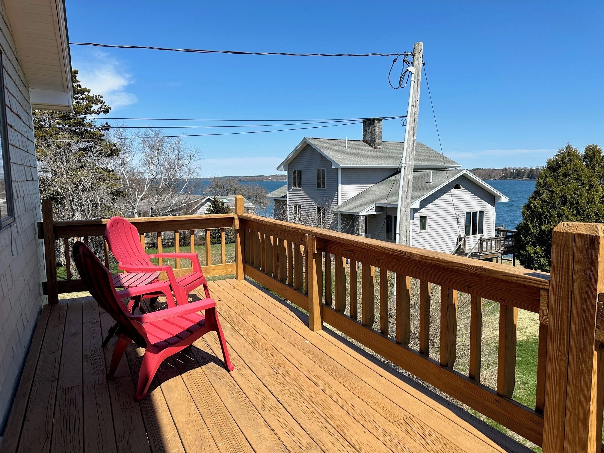 Une terrasse en bois avec une rambarde, une chaise rouge et une vue sur un lac et des maisons au loin.