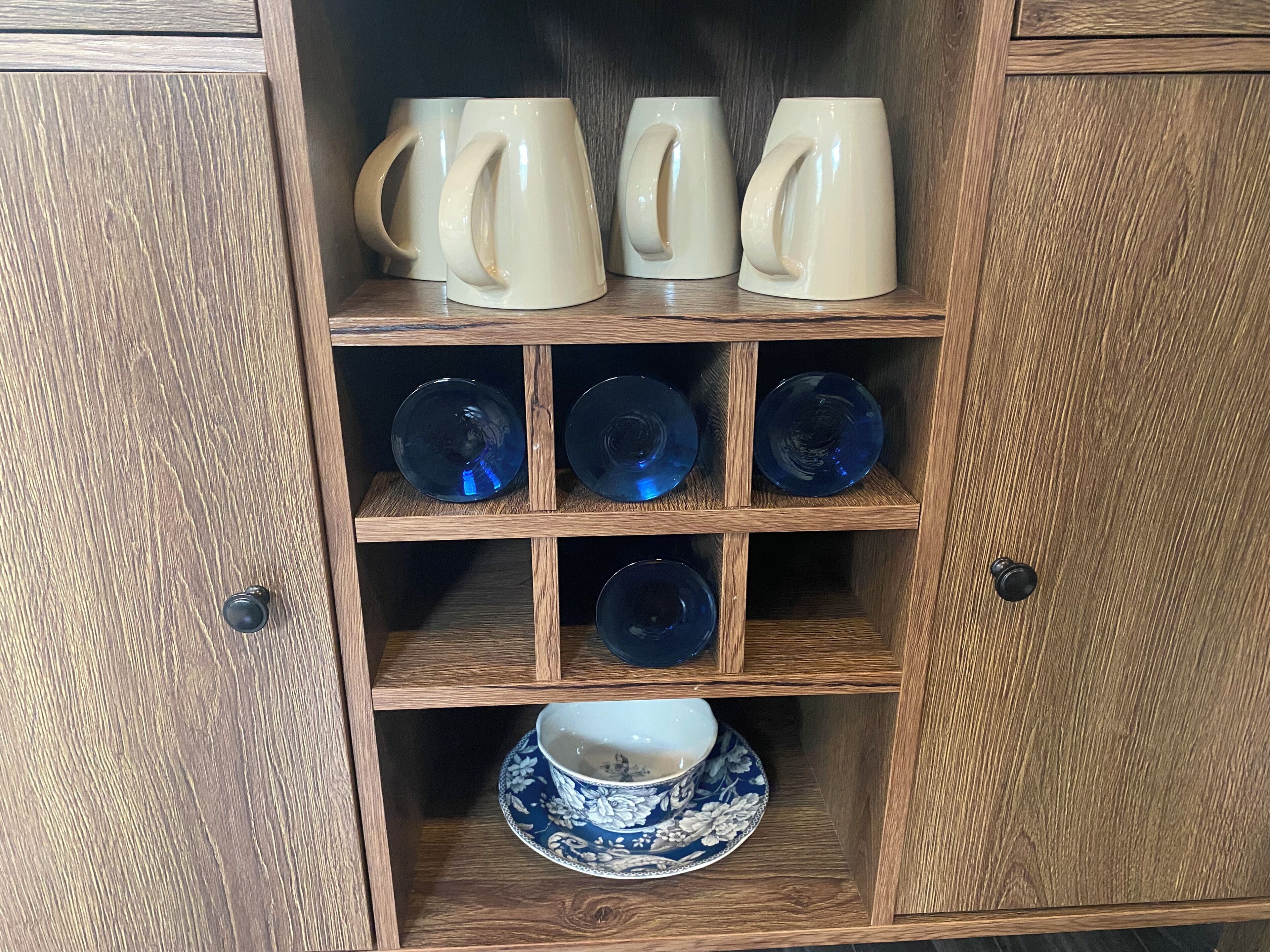 A wooden cabinet with shelves holding ceramic mugs, blue glass bowls, and a blue and white floral bowl set.
