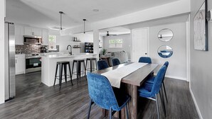 A modern kitchen with a central island, bar stools, and a dining area with a long table and blue chairs. - 1.4 Mi to AT&T | Mini Golf + Hoops | Sleeps 12 (Arlington)