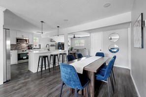 A modern kitchen with a central island, bar stools, and a dining area with a long table and blue chairs. - 1.4 Mi to AT&T | Mini Golf + Hoops | Sleeps 12 (Arlington)