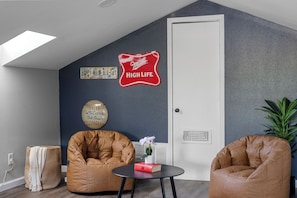 A room with a high ceiling, a door, two bean bag chairs, a small table with a book and a vase, and wall decorations including a baseball and a Miller High Life sign.