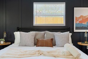 A bedroom with a bed, a window with blinds, a framed artwork, and bedside tables with lamps.