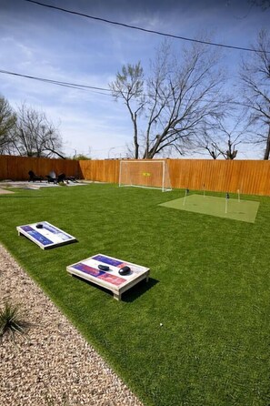 A backyard with artificial grass, two shuffleboard tables, a wooden fence, and a soccer goal. - Sleeps 12 | Pickleball + Mini Golf + Pool Table (Arlington)