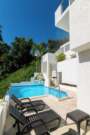 A swimming pool with lounge chairs and a small table, surrounded by a white building with a staircase and a view of greenery.