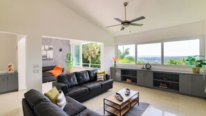 A modern living room with a black leather sofa, a coffee table, and a view of palm trees through large windows. - Casa Mango- Relaxing Oasis -10 mins drive to the beach, EPIC views & pool (Puerto Plata)