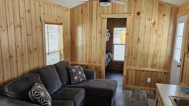 A cozy cabin interior with a wooden exterior, a sofa with patterned cushions, a dining area with a sink, and a ceiling fan. - Relax in the Fox Den | Private 2BR Cabin Close to Beach & Activities (Isle)