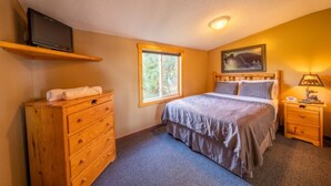 Un dormitorio con cama, cómoda, mesita de noche, televisor en un estante, ventana y un cuadro en la pared.