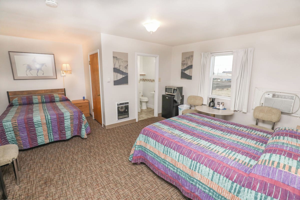 A hotel room with two beds, a desk, a chair, a microwave, a refrigerator, and a window with curtains.