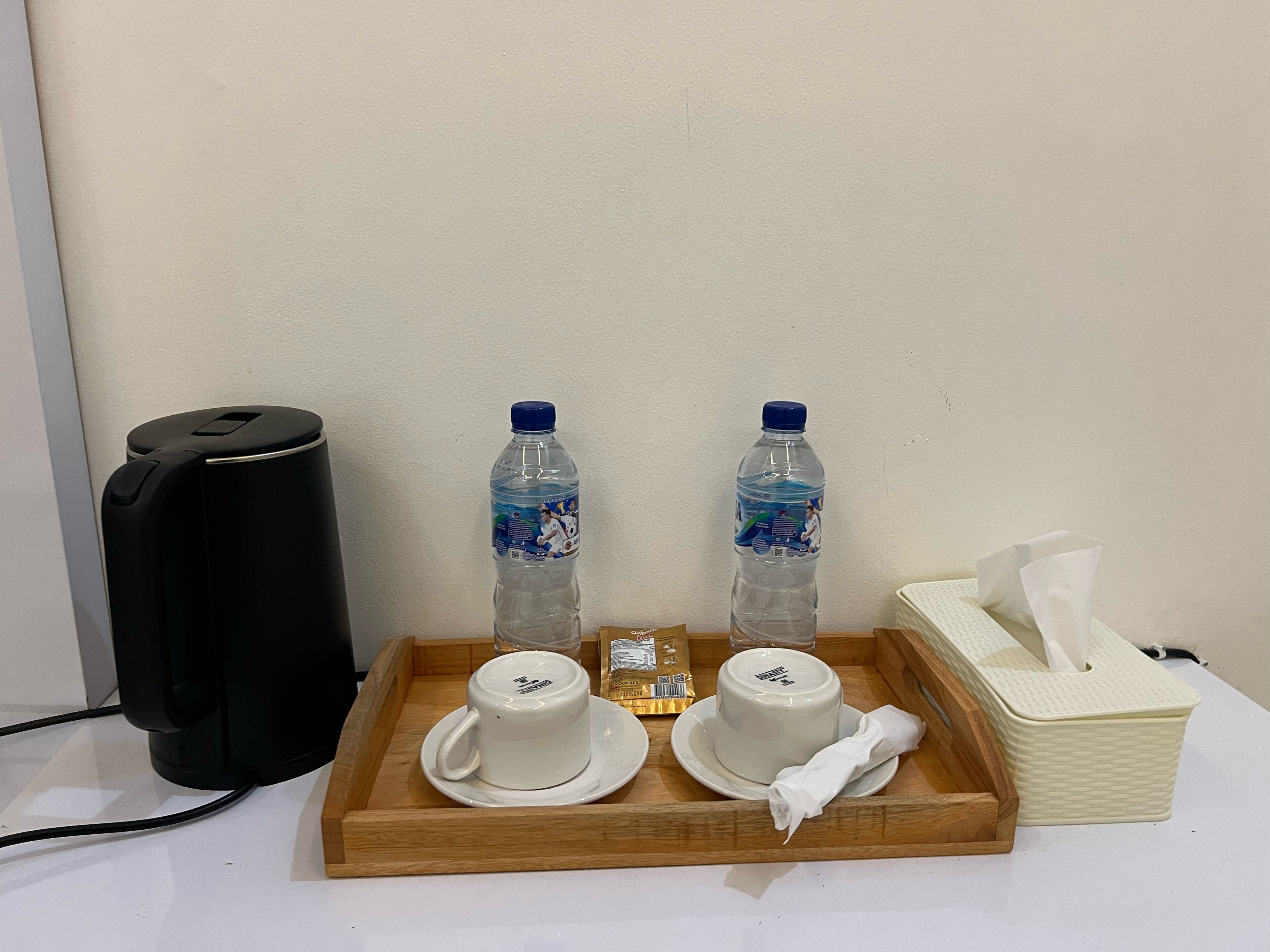 Una bandeja con dos tazas y platillos blancos, dos botellas de agua, una caja de pañuelos y un dispositivo negro sobre una bandeja de madera.