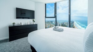 Une chambre moderne avec un grand lit, une commode noire, un téléviseur et une vue sur la ville grâce à de grandes fenêtres.
