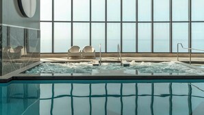 Une grande piscine intérieure avec de larges baies vitrées donnant sur la mer. On aperçoit deux chaises longues et un jacuzzi au fond.