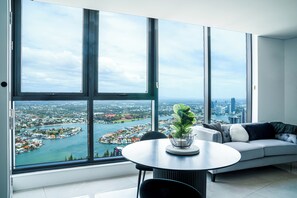 A modern living room with a large window, a round table, and a grey sofa with cushions. - Breathtaking Ocean Views 1BD+Amazing Facilities! (Surfers Paradise)