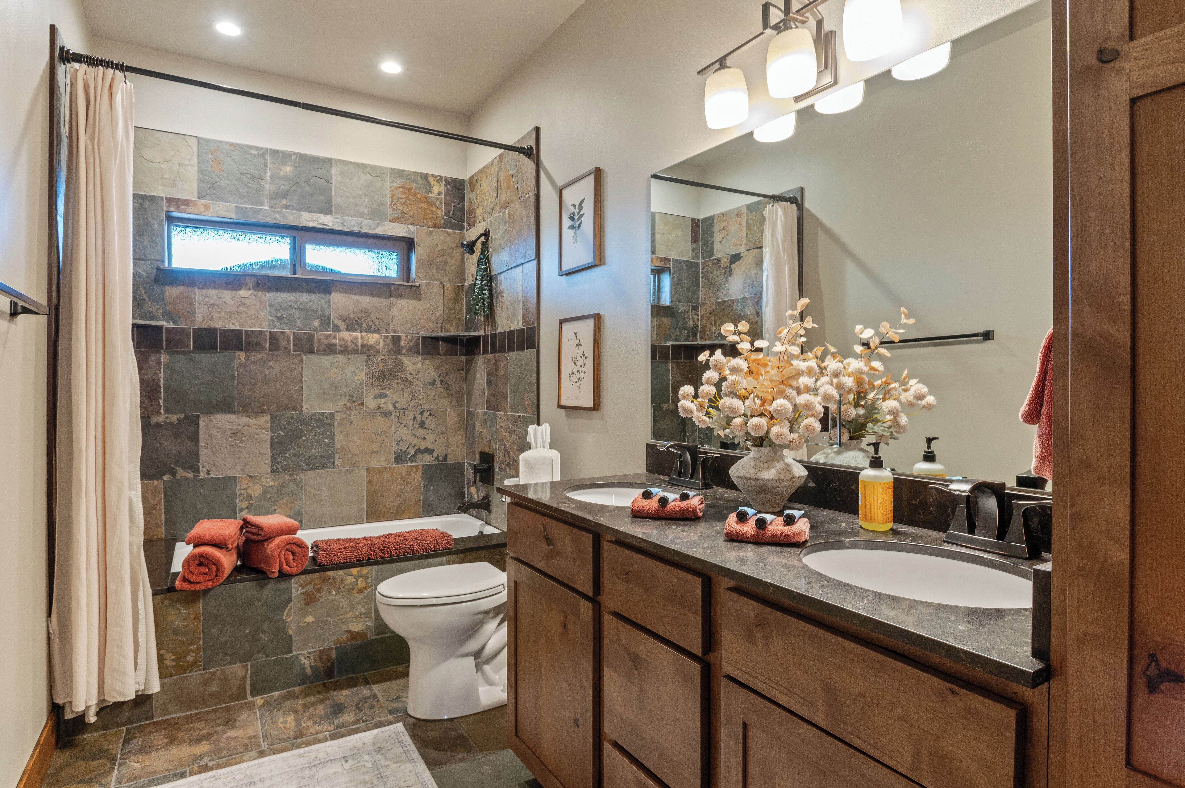 A bathroom with a stone shower wall, a white toilet, a vanity with two sinks, and a large mirror.