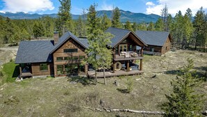 A modern log cabin with a large deck and a second-story balcony, surrounded by a forest and mountains.
