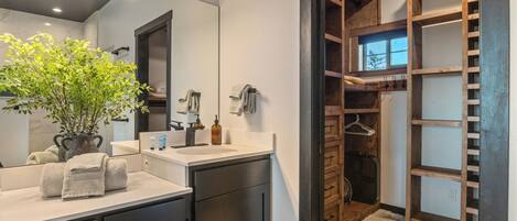 A modern bathroom with a large mirror, a sink with a towel, a plant, and a wooden storage unit.