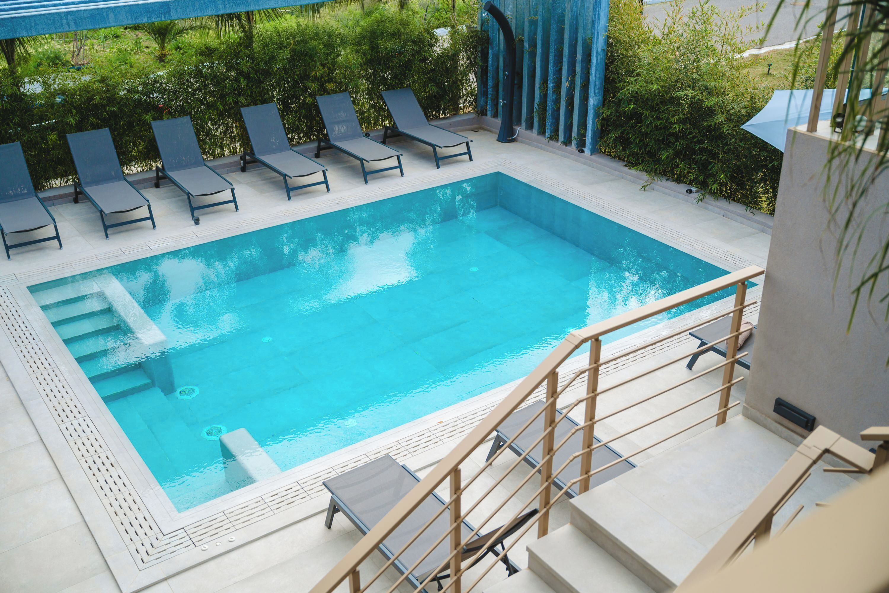 Ein rechteckiger Pool mit einer gefliesten Liegefläche, umgeben von Liegestühlen und einer hölzernen Treppe, die hinunterführt.