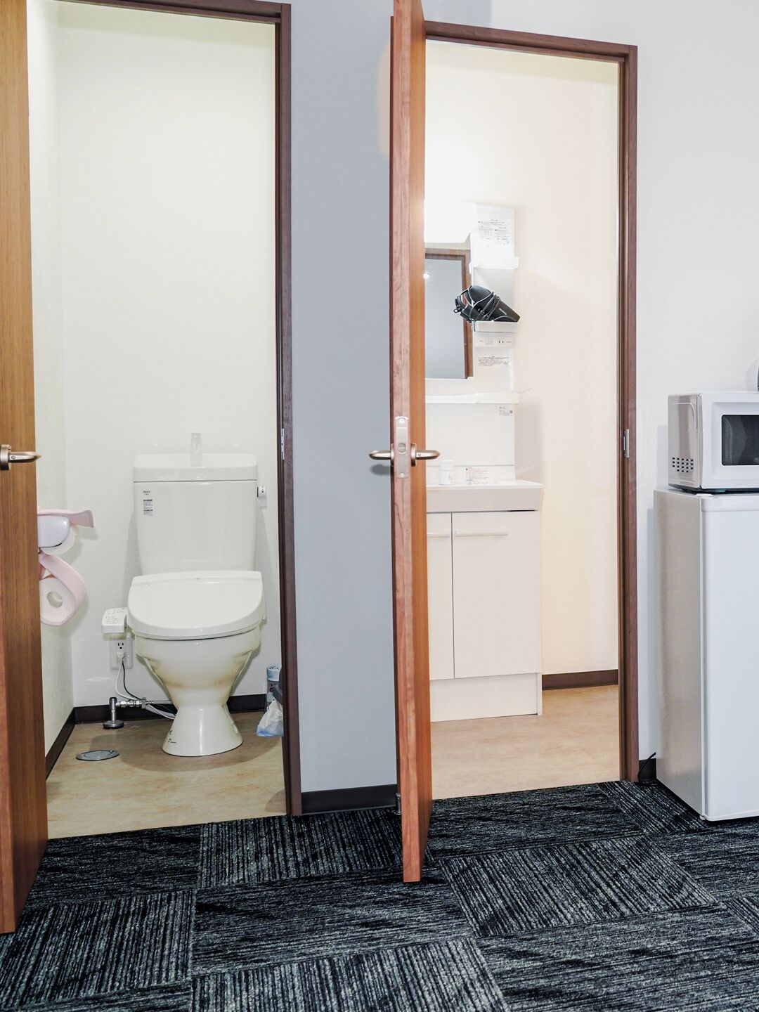 Habitación de hotel con inodoro, lavamanos, microondas y una puerta que da a otra habitación.