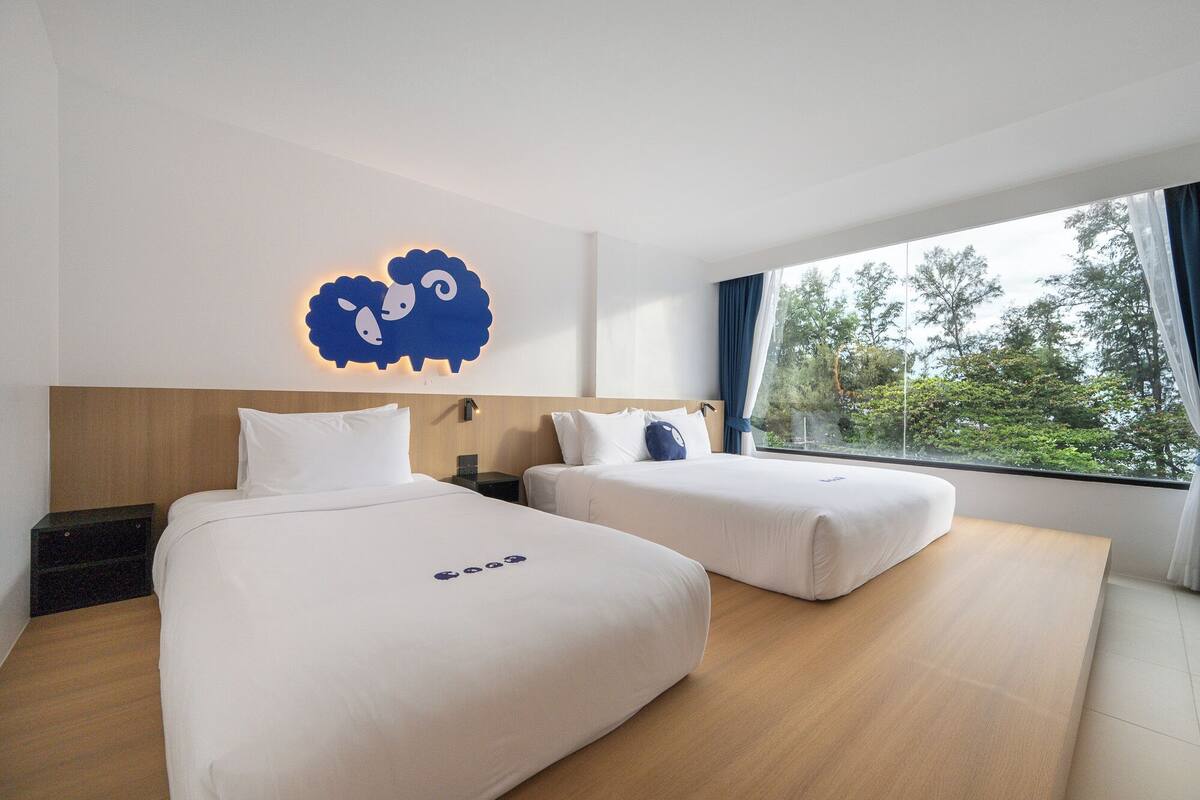 A hotel room with two beds, a wooden headboard, a wall-mounted light fixture, and a window with a view of trees.