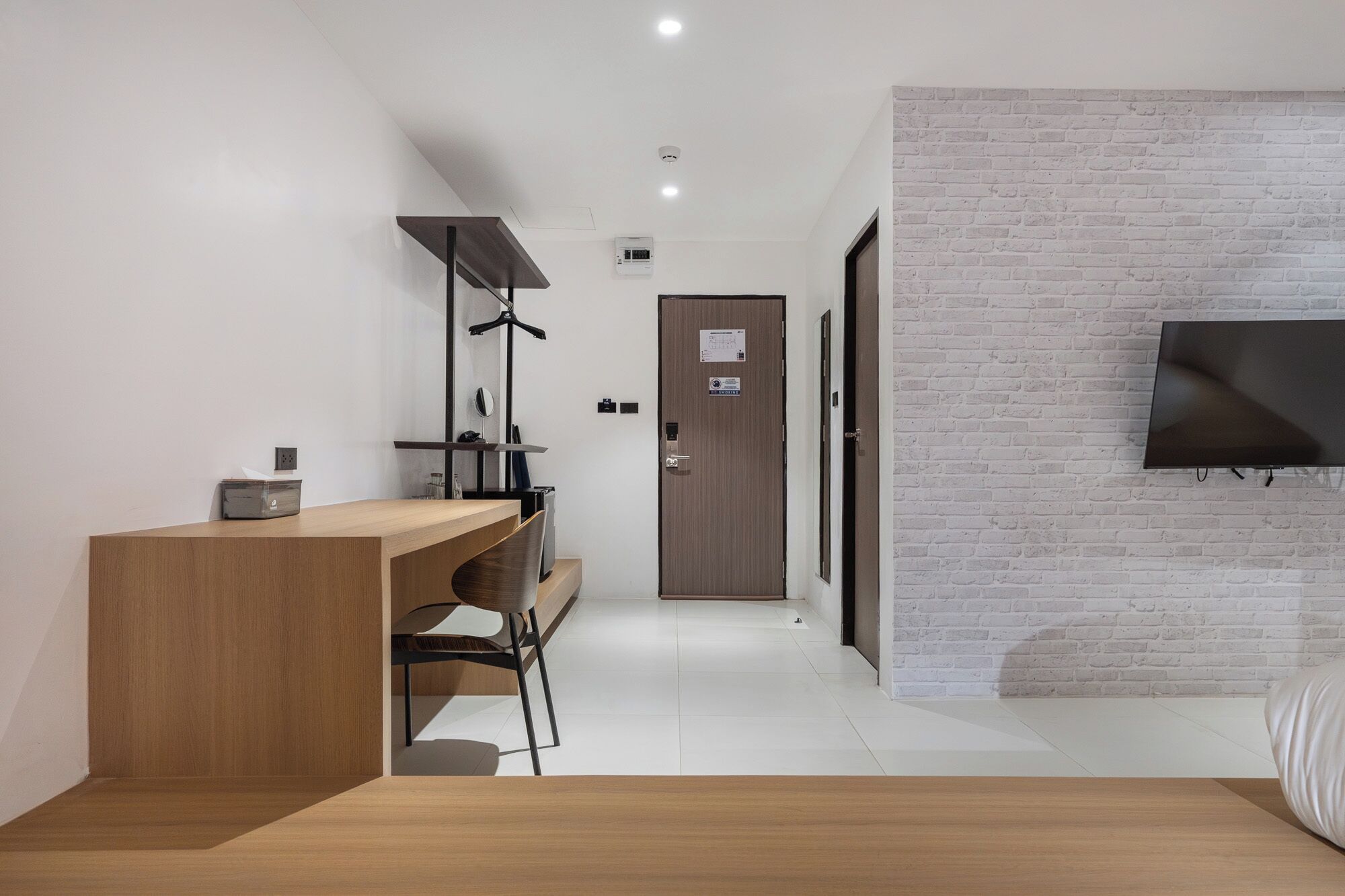 A modern hotel room with a wooden desk, a chair, a flat-screen TV mounted on the wall, and a white brick accent wall.