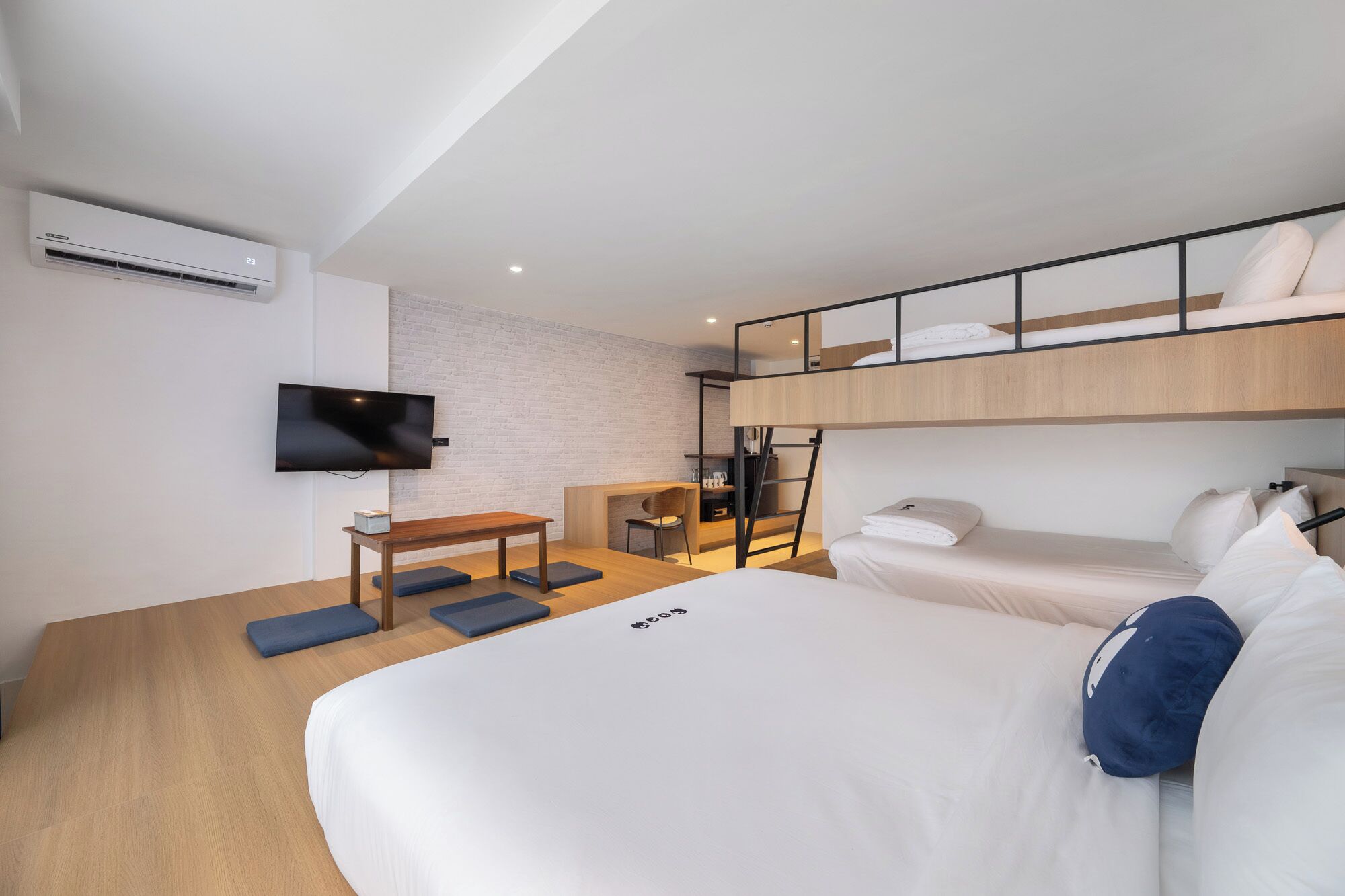 A modern hotel room with a bunk bed, a desk with a chair, a flat-screen TV mounted on the wall, and a small seating area with cushions.