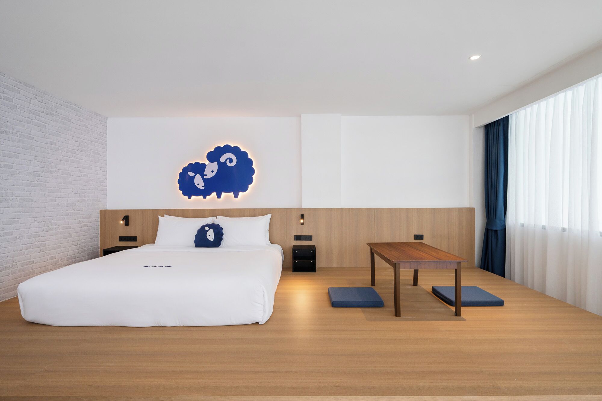 A modern hotel room with a large bed, a wooden nightstand, a small table, and a wall-mounted light fixture.