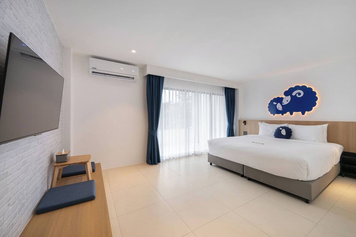 A modern hotel room with a large bed, a flat-screen TV, a wooden desk, and a large window with curtains.