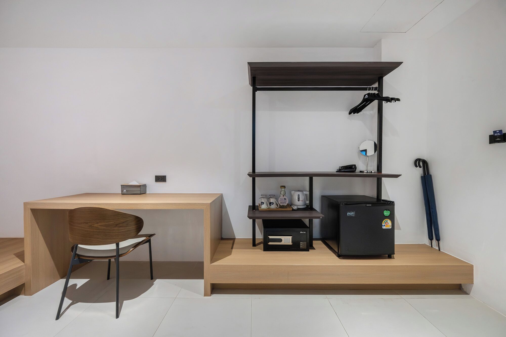 A compact, modern room with a wooden desk, a chair, a small black refrigerator, and a shelf unit with various items.