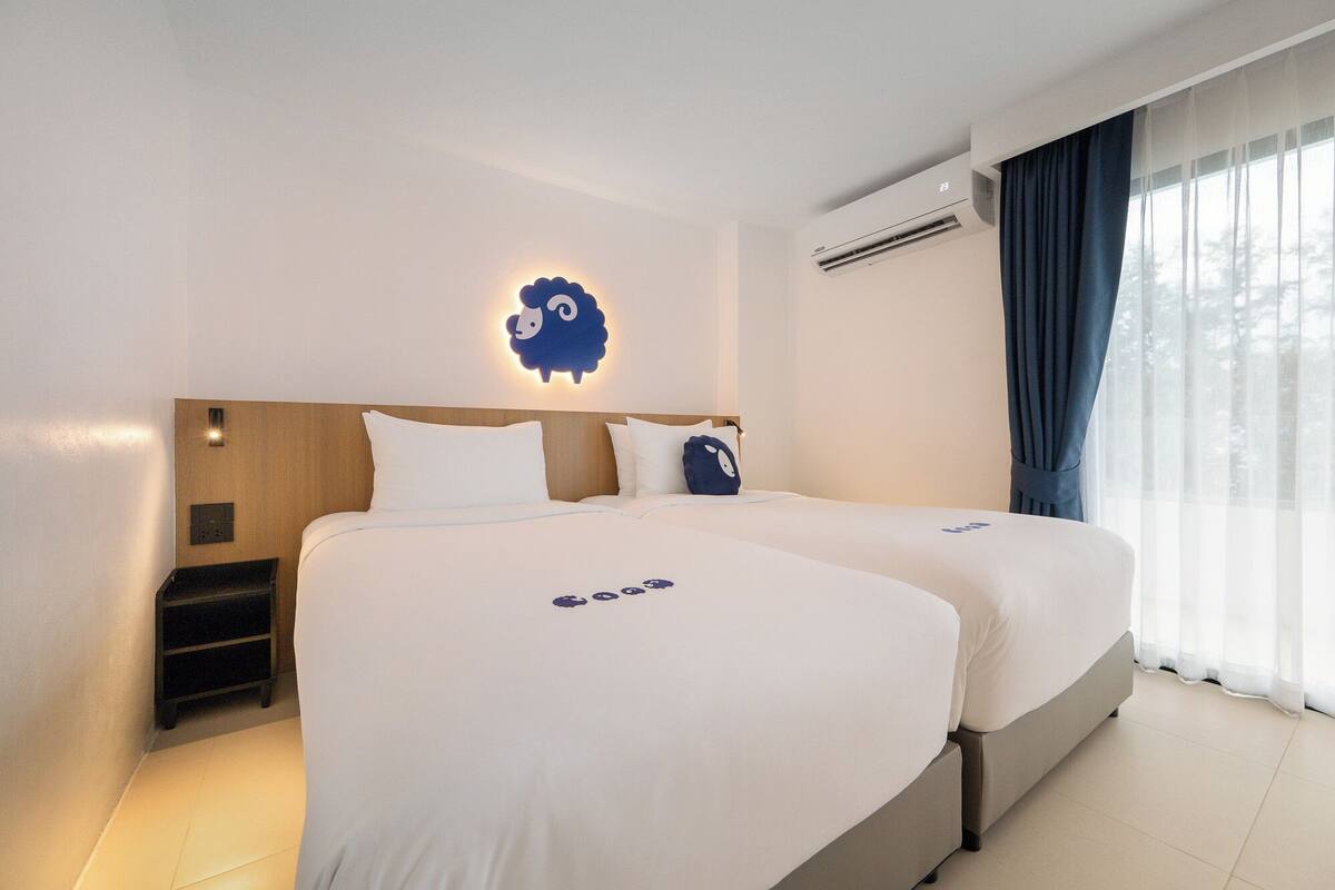 A hotel room with two beds, a wall-mounted air conditioner, a nightstand, and a window with curtains.