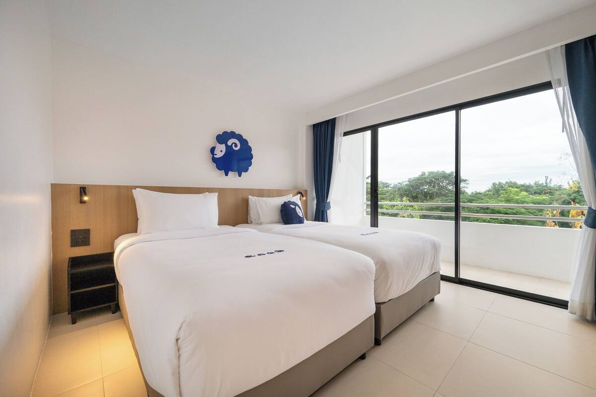A hotel room with two beds, a wooden headboard, a large window with a view of greenery, and a blue decorative wall piece.
