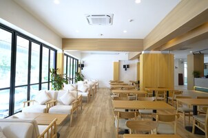Un espacio interior amplio con varias mesas de madera y asientos acolchados blancos, ventanas grandes y un equipo de aire acondicionado instalado en el techo.