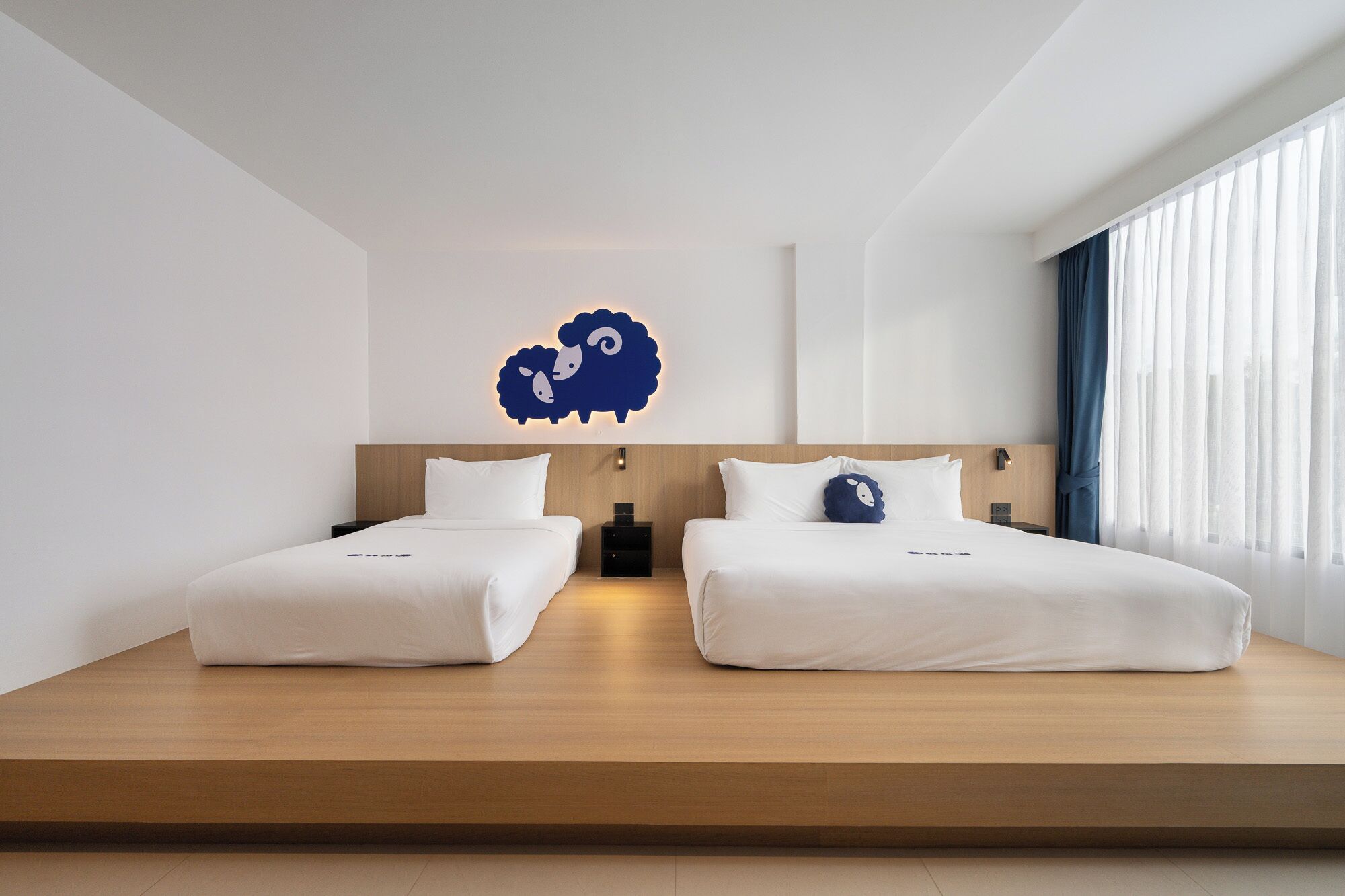 Une chambre d’hôtel moderne avec deux lits, une plateforme en bois et une décoration murale représentant un mouton.