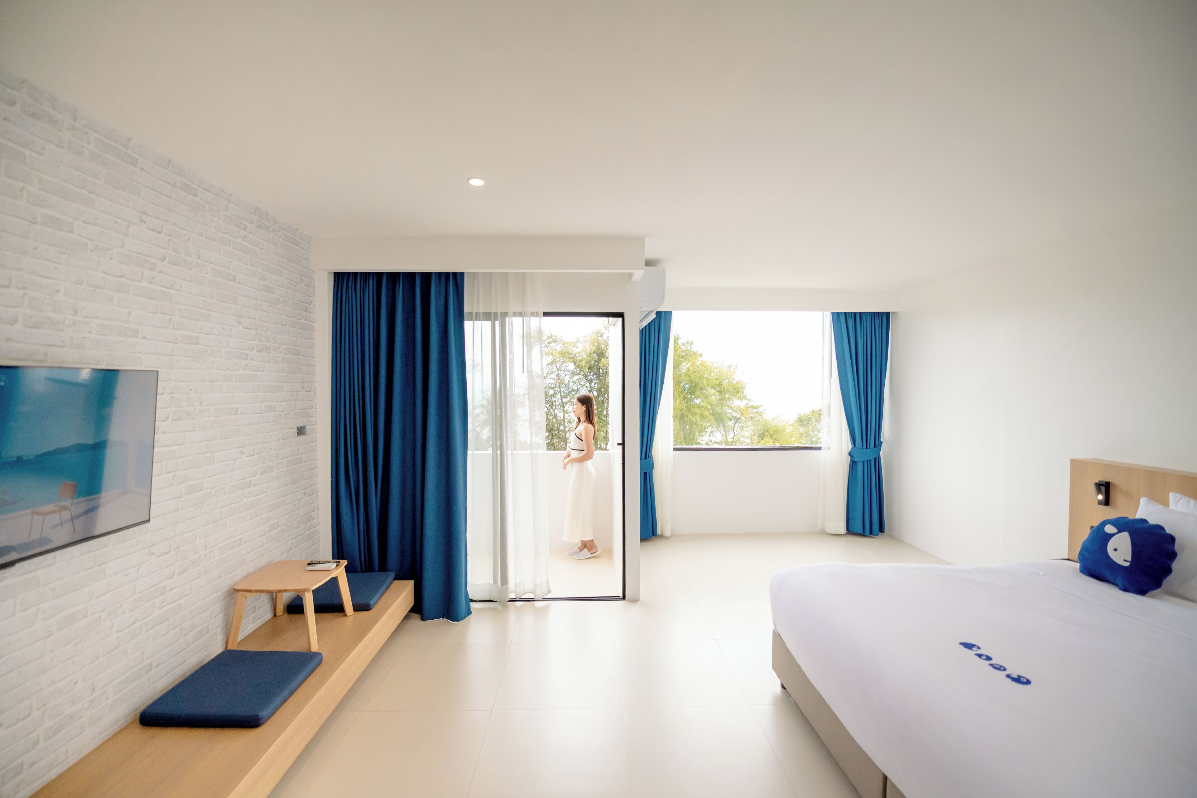 A modern hotel room with a white brick wall, a bed with a blue pillow, a small wooden table, a bench, and a TV mounted on the wall.