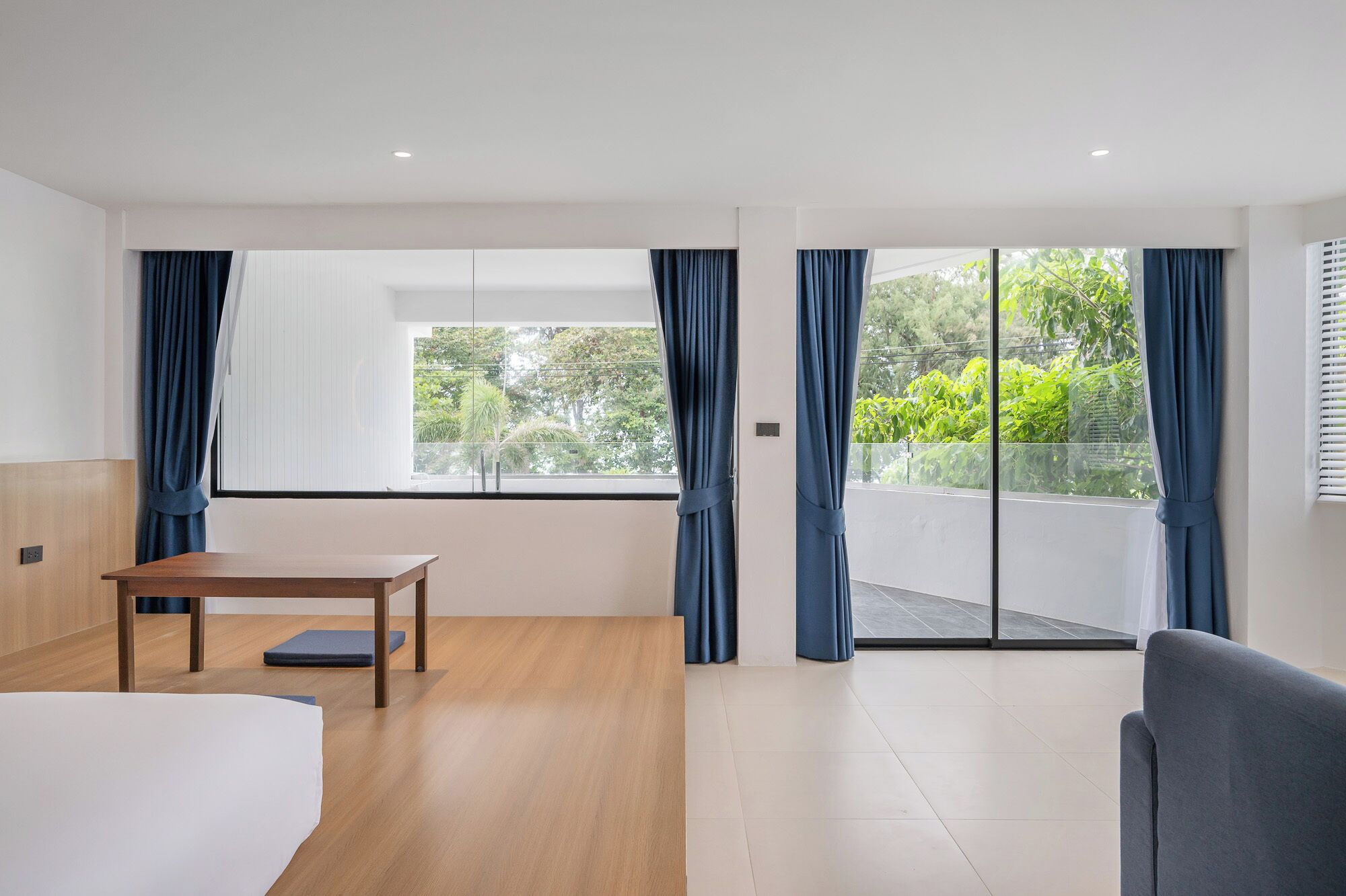 Una habitación de hotel moderna con un amplio ventanal corredera que da a un balcón, una mesa de madera y una cama con ropa blanca.