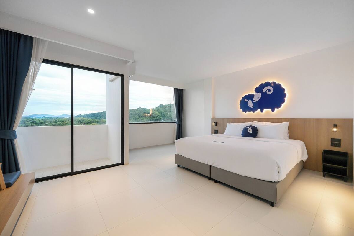 A modern hotel room with a large bed, a sliding glass door to a balcony, and a decorative sheep wall art piece.