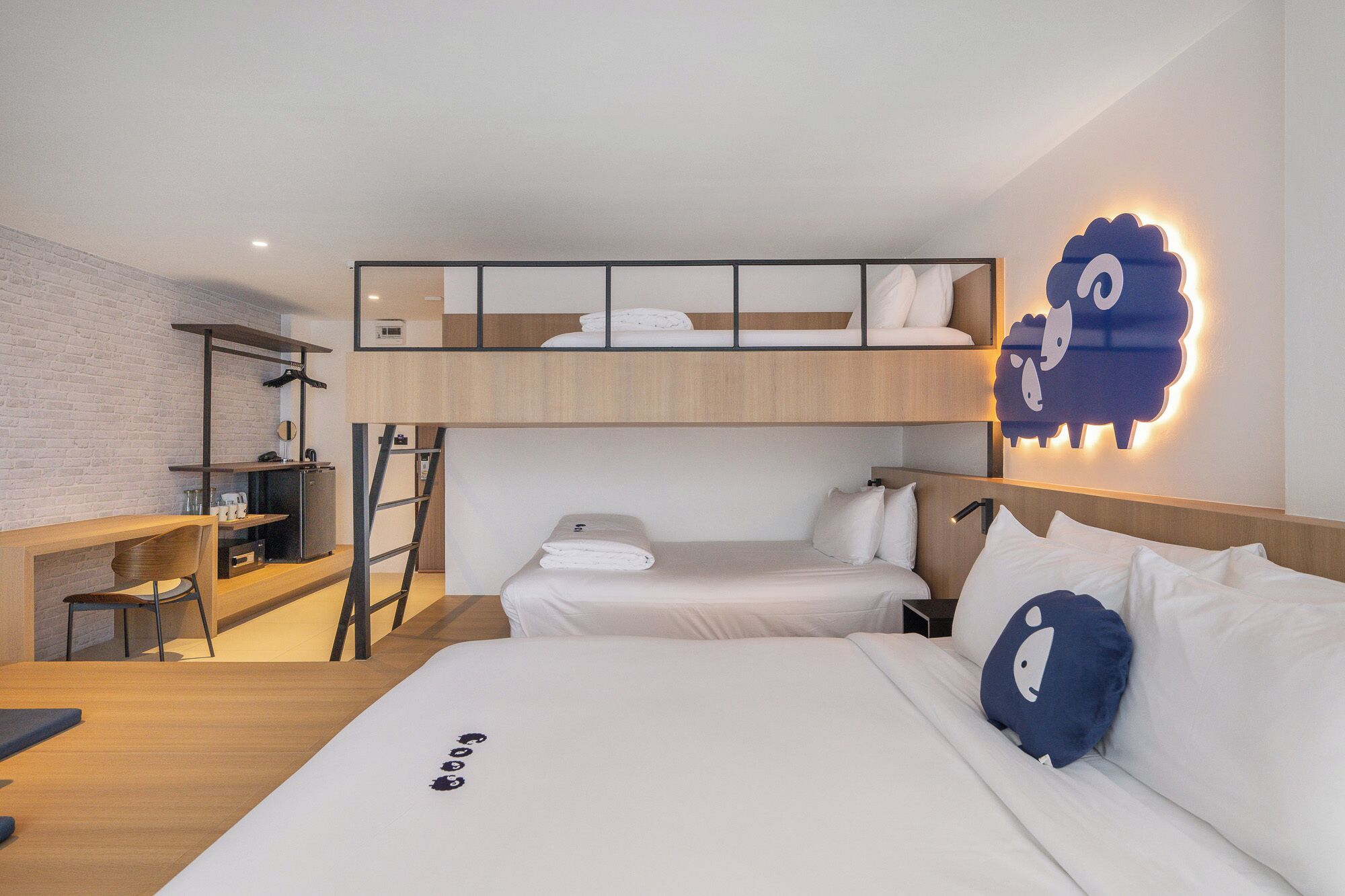 Una habitación de hotel moderna con una cama litera, un escritorio con silla, una kitchenette pequeña y una pieza decorativa de arte mural con un borrego.