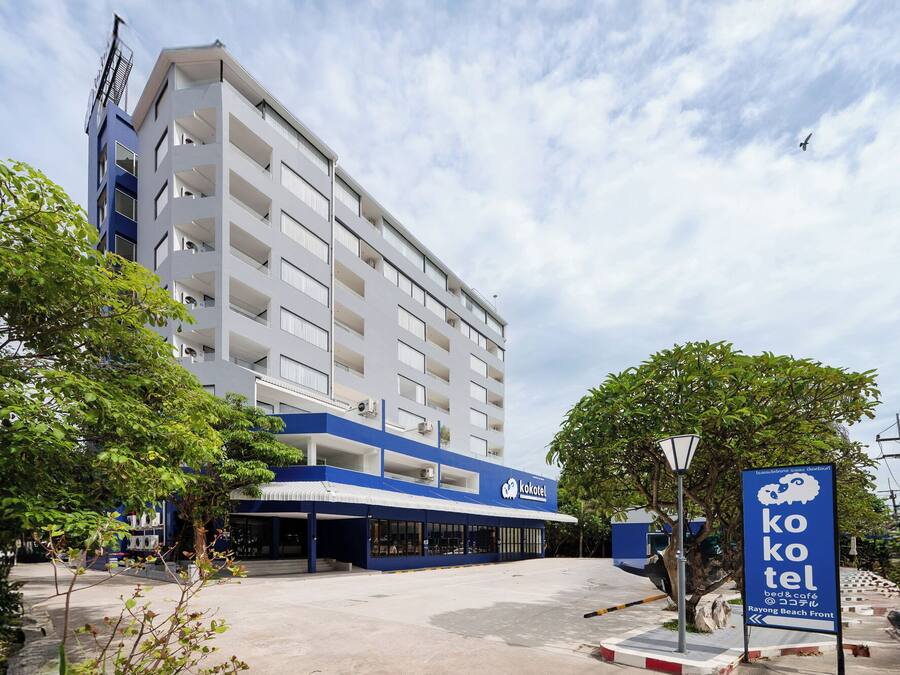 Un edificio moderno con fachada azul y blanca, un letrero que dice 'kokotel' indicando una posada, y vista al frente de la playa Rayong.